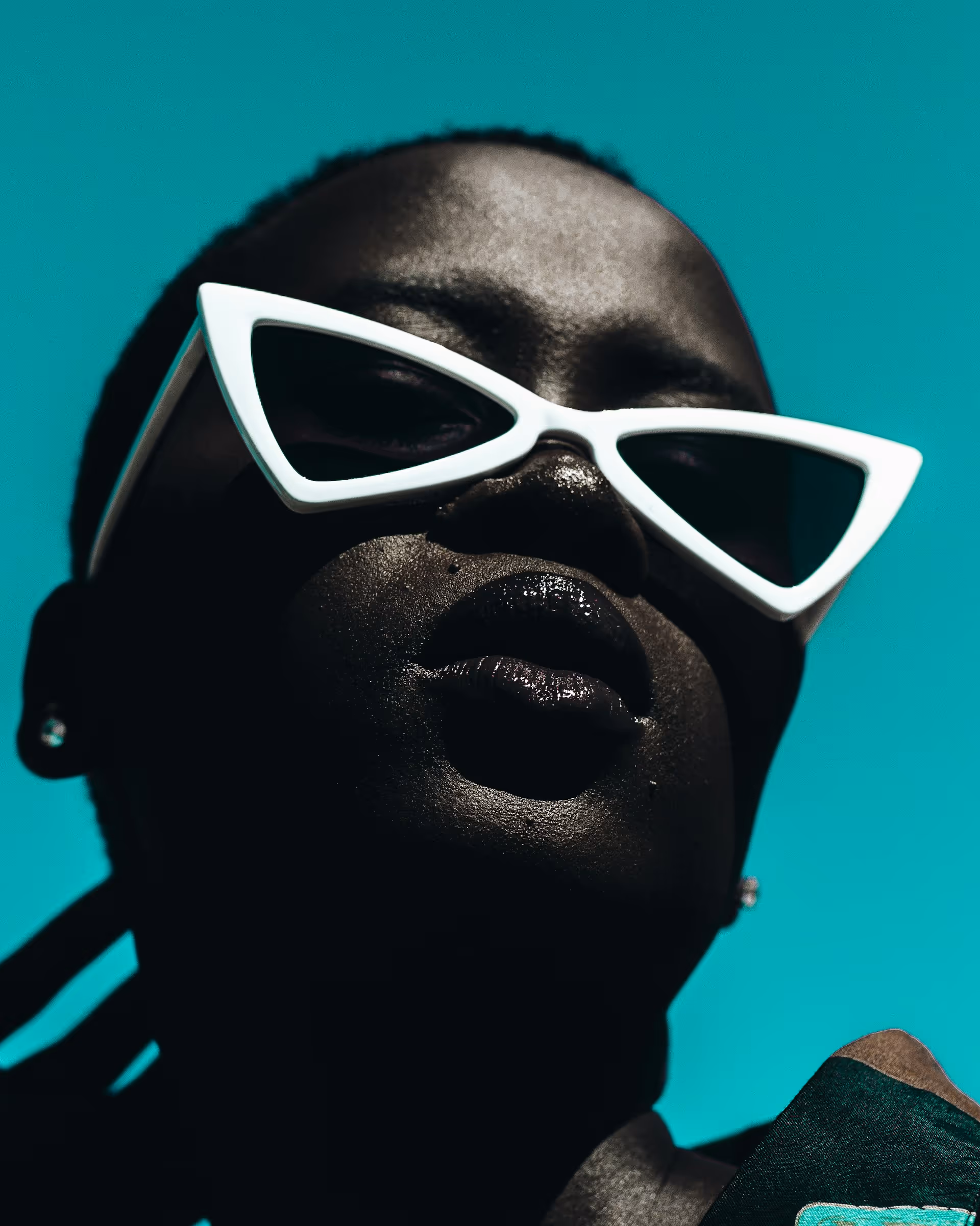 Close-up of a person with dark skin wearing white cat-eye sunglasses against a turquoise background.
