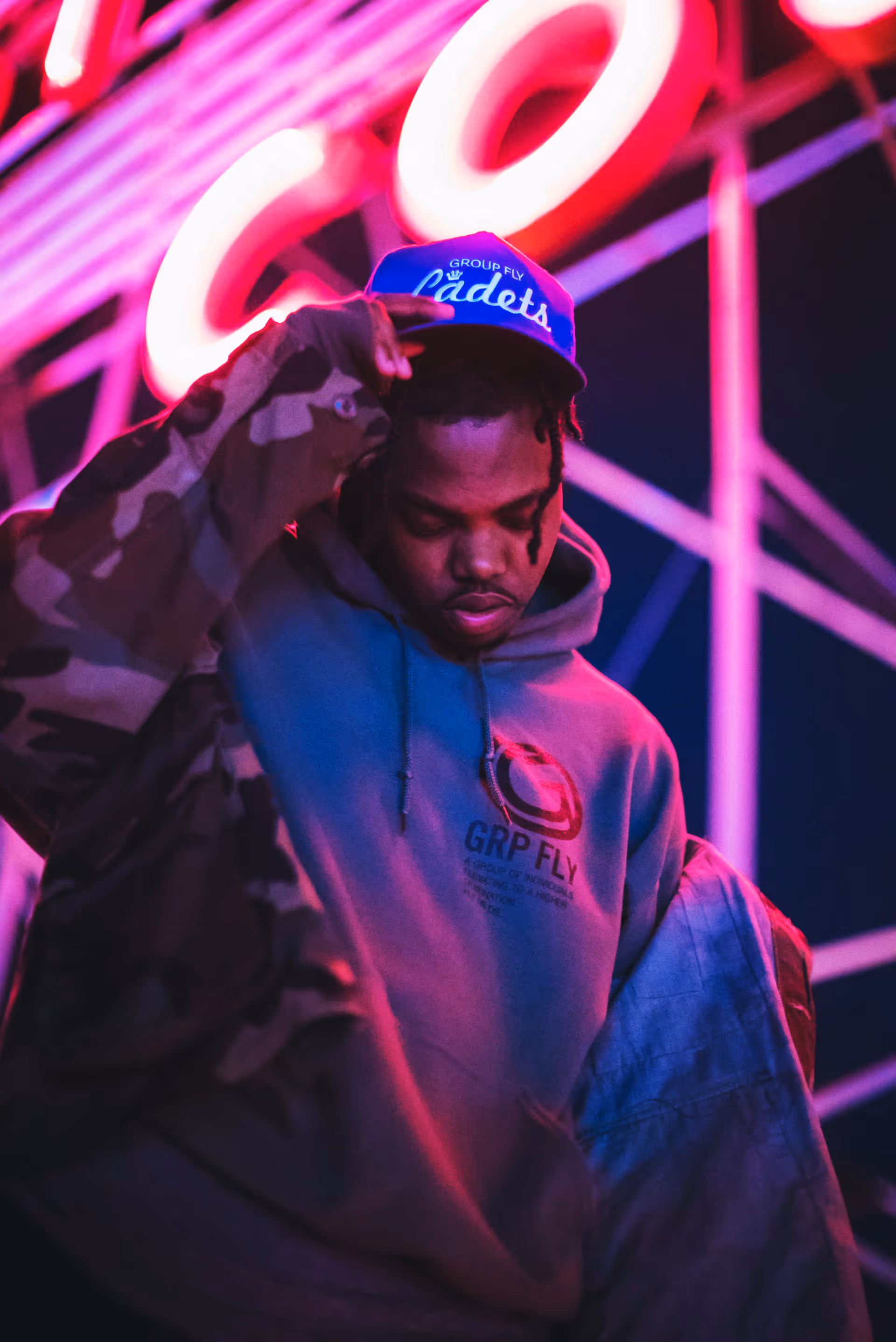 Man wearing a blue cap and grey hoodie with GRP FLY logo, posing under pink neon lights.